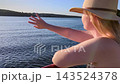 A young woman in a straw hat and sundress enjoys a ferry boat cruise on the water during a scenic sunset. She stands on the deck enjoying peaceful travel and golden hour light. 143524378