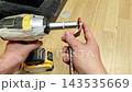 Impact driver and red bracket stay aligned as fastening continues. Hands hold metal part in place while threaded rod extends outward, creating precise tool adjustment task on workbench. 143535669