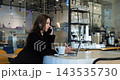 serious young woman conducts remote work conversation on mobile phone while browsing laptop inside contemporary coffee shop, sitting at high table with drink during business discussion. 143535730