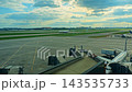Passenger aircraft rests beside boarding bridge while distant airplane glides smoothly along runway tarmac. Airport terminal view showcases parked airliners preparing for international flight. 143535733