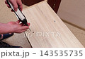 Hands use power driver on wood panel for interior project. Screws are set into predrilled holes on floor, creating neat assembly step with careful attention and purposeful movement. 143535735