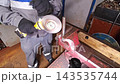 Gloved worker operates yellow angle grinder on clamped metal stock inside workshop. Metal grinding happens beside red vise and wooden block, creating practical repair and fabrication task. 143535744