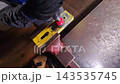Worker drills through yellow steel plate with power tool during workshop metalwork. Drilling action stays focused on clamped workpiece, with gloved hands controlling pressure and alignment. 143535745