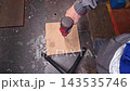 Wood drilling in workshop as gloved hands guide power drill into board. Worker steadies wood on workbench, creating clean hole during careful drilling practice with focused effort. 143535746