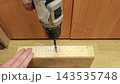 Wood drill operation continues on timber board as hand holds material steady. Power drill makes precise hole in workshop lighting, with focused manual control and practical carpentry use. 143535748