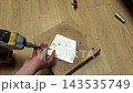 Impact driver fastens metal plate during assembly work. Hands guide screw into board while power tool tightens joint, with careful focus on precise placement and secure attachment. 143535749