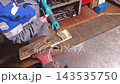 Jigsaw cutting wood in workshop. Worker holds power saw against secured plank while vise keeps material steady, continuing careful bench task with visible concentration and practical tool use. 143535750
