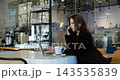 Focused businesswoman talks on phone and works on computer at cafe table, maintaining concentrated discussion. Professional woman handles remote workplace duties beside cup of hot coffee. 143535839