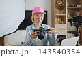 A smiling caucasian woman in a pink cap holds a camera and prepares to record content in her home studio with lighting and equipment 143540354