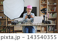 A young Caucasian woman wearing a pink cap and denim shirt is examining a digital camera at her desk, surrounded by photography equipment and a laptop 143540355