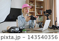 A smiling caucasian woman wearing a pink cap holds a professional camera while surrounded by photography equipment in her home studio 143540356