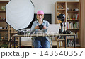A woman with a pink cap sits at a desk surrounded by photography equipment, including cameras, a laptop, and a softbox light 143540357