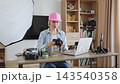 A young woman wearing a pink cap and denim shirt is reviewing footage on her camera in a home studio setup with lighting and equipment 143540358