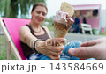 Woman sits in a lounge chair and smiles giving an ice cream cone to her partner on a sunny day at a ice cream food truck 143584669