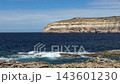 Limestone cliffs at Dwejra Bay on Gozo island in Malta rise above the Mediterranean Sea as waves break on a rocky shore. Coastal landscape, scenic travel, island sightseeing, natural rock formations. 143601230