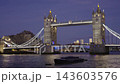 Static medium shot of floodlit Tower Bridge over the River Thames during blue hour twilight, featuring red London buses crossing the deck. 143603576