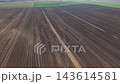 Aerial drone view reveals tractor tire tracks crossing freshly plowed agricultural field in rural Germany. Curving wheel marks intersect parallel soil furrows, creating abstract geometric patterns 143614581