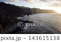 A drone flies low along the sunlit cliffs of the Anaga coast near Benijo with the ocean glowing under a low sun. Dramatic backlit island scenery for travel productions. 143615138