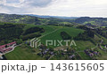 Drone perspective over wide green countryside with rolling hills, patchwork fields, and winding roads leading past farm buildings to distant ridges under soft cloudy daylight. 143615605