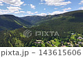 Overhead perspective reveals deep mountain basin covered in evergreen trees, village homes along valley floor, winding roads, and bright sunlit meadows under clear blue sky with clouds. 143615606