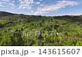 Wide aerial perspective shows quiet rural hillside village with clustered homes and tall church spire, bordered by green fields, orchards, winding roads. 143615607