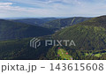 Wide aerial view across mountain valley, showing dense forest ridges, scattered villages, and winding roads weaving through green terrain and open meadows. 143615608