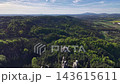 Aerial panorama shows dense forest opening into rolling valley, rocky outcrops, patchwork farmland, and small distant town beneath bright clear sky, conveying calm rural scale. 143615611