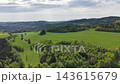 High-altitude aerial view shows patchwork fields and grassy lanes bordered by dense tree belts. Sunlit clouds create soft highlights and gentle shadows across rolling hills, clearings. 143615679
