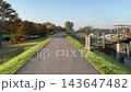 秋の朝、紅葉した木々が並ぶ河川敷の舗装された遊歩道を歩く風景 143647482
