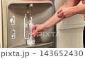 A person stands at a water refill station and fills a plastic bottle with water. The sun is out and the area is busy 143652430