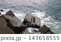 Ocean waves crash against massive concrete tetrapods protecting a rugged coastline, creating swirling turquoise water and white foam. 143658555