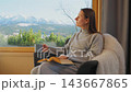 A woman reading by a large window and enjoying scenic snowy mountain view. 143667865