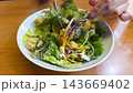 Tossing a fresh garden salad in a white bowl on a wooden table. The mix includes leafy greens, shredded carrots, croutons, and radish with a creamy dressing. 143669402