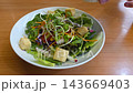 A person pours creamy dressing over a fresh garden salad in a white bowl, containing mixed greens, croutons, sprouts, and carrots, set on a wooden table. 143669403