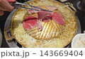 Thin slices of meat cook on a traditional Thai barbecue dome surrounded by simmering broth and cabbage. Hands use tongs and chopsticks to grill the food during a communal meal. 143669404