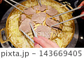 Chopsticks lift a thin slice of meat from a traditional brass Thai barbecue pan, or mu kratha, featuring a central dome for grilling and a trough of simmering broth with cabbage. 143669405