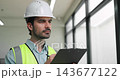 Portrait of Hispanic engineer reviews clipboard near tall windows during building inspection. Focused construction worker writes project notes checking form 143677122