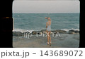 Girl in a swimsuit stands on a rocky shore looking at ocean waves, captured in slow motion on vintage Super 8 film 143689072