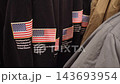 Many hoodies with sewn rubber flags on sleeve of black fleece sweatshirt hang on rack in store. 3D patches decorate branded American national apparel in clothing shop. 143693954