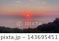 Aerial view of bright red sun setting over calm sea horizon with glowing light reflection on water and dark trees in foreground for sunset video footage. 143695451