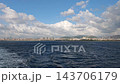 Wide view of a distant urban skyline across rippling water, filmed from a moving boat under a blue sky with large clouds. 143706179