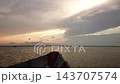 Wooden boat bow gliding across calm lake at sunset under layered dramatic clouds, golden pink sky and distant shoreline visible in soft evening light, peaceful forward motion from low viewpoint 143707574