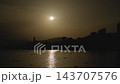 Partially eclipsed sun over silhouetted city skyline with shimmering water reflection and swimmers at dusk, warm golden backlight on calm waterfront scene, distant hills and high-rise outlines visible 143707576
