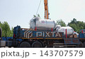 Crane lifting cylindrical stainless steel storage tanks wrapped in protective plastic onto flatbed truck at outdoor construction site, bright midday sky with trees in background 143707579
