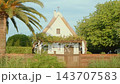 Small white chapel with steep thatched roof, cross, vine-covered pergola, and wooden gate framed by palm and dense greenery in warm golden light, frontal composition for tranquil rural scene. 143707583