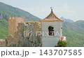 Sunlit stone bell tower with terracotta tiled roof on cliff beside medieval fortress ruins overlooking pine covered mountain valley and distant peaks from elevated viewpoint, daytime scenic landscape, 143707585