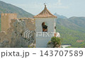 Whitewashed bell tower with terracotta roof perched on rocky cliff above pine valley in sunlit mountain landscape, bell visible through arched opening, serene travel and heritage scene 143707589