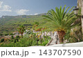 Palm tree lined hillside promenade viewed through rustic stone archway on sunny day, white plaster foreground and winding pedestrian path leading into lush green valley, distant mountains under blue 143707590