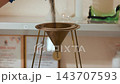 Technician pouring dark granular powder through metal cylinder into brass funnel on tripod over laboratory bench, closeup of gloved hand and steady stream of material being transferred 143707593