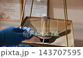 Close-up of metallic cylinder topped with dark powder being sampled with glass slide inside brass laboratory stand under warm light while gloved hand holds slide for inspection. 143707595